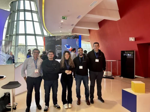 Group of people posing together in a modern venue during a Deucalion-related event, with a large screen displaying technical content in the background and natural light from a glass wall.
