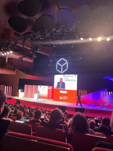 Audience seated in a conference hall watching a speaker on stage at an event focused on quantum and AI communities, with large screens and stage lighting.