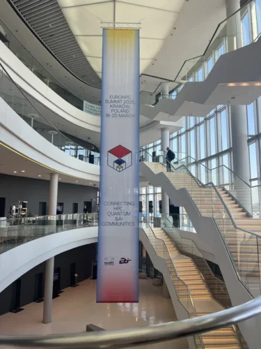Interior of a modern conference venue with a large vertical banner for the EuroHPC Summit 2025 in Kraków, featuring staircases, glass walls, and open architectural design.
