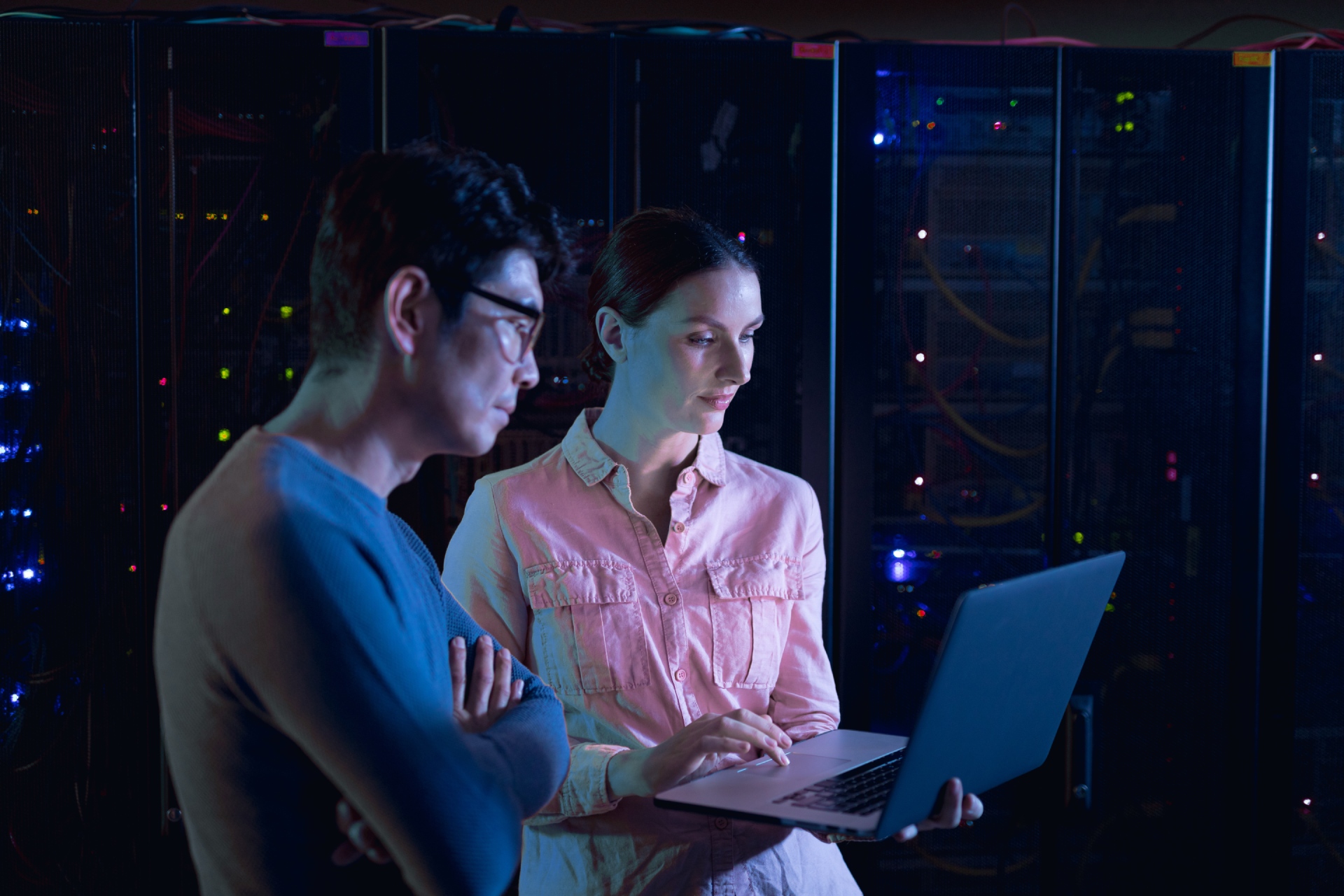 Two IT professionals standing in a server room, reviewing information on a laptop while surrounded by illuminated server racks.