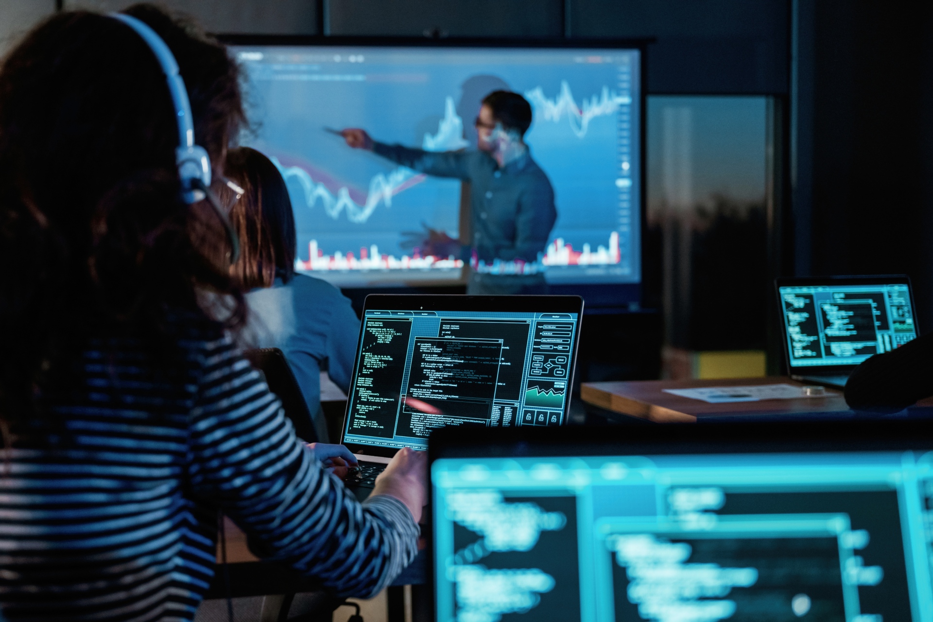 People working on laptops in a dark classroom or training room while an instructor presents charts and data on a large screen at the front.
