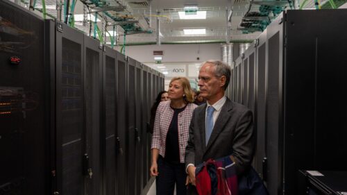 Delegation walking through Deucalion data centre aisle, surrounded by HPC server racks and cooling systems.