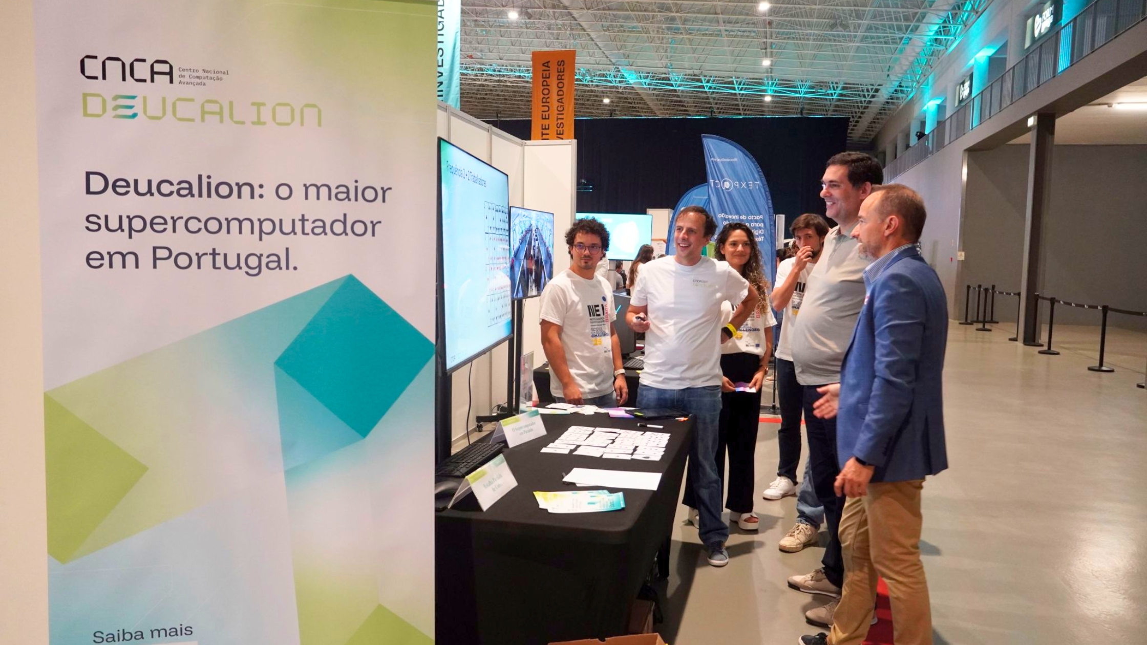 People gathered around a Deucalion information stand at an event venue, with a banner reading “Deucalion: o maior supercomputador em Portugal” and informational materials displayed on a table.