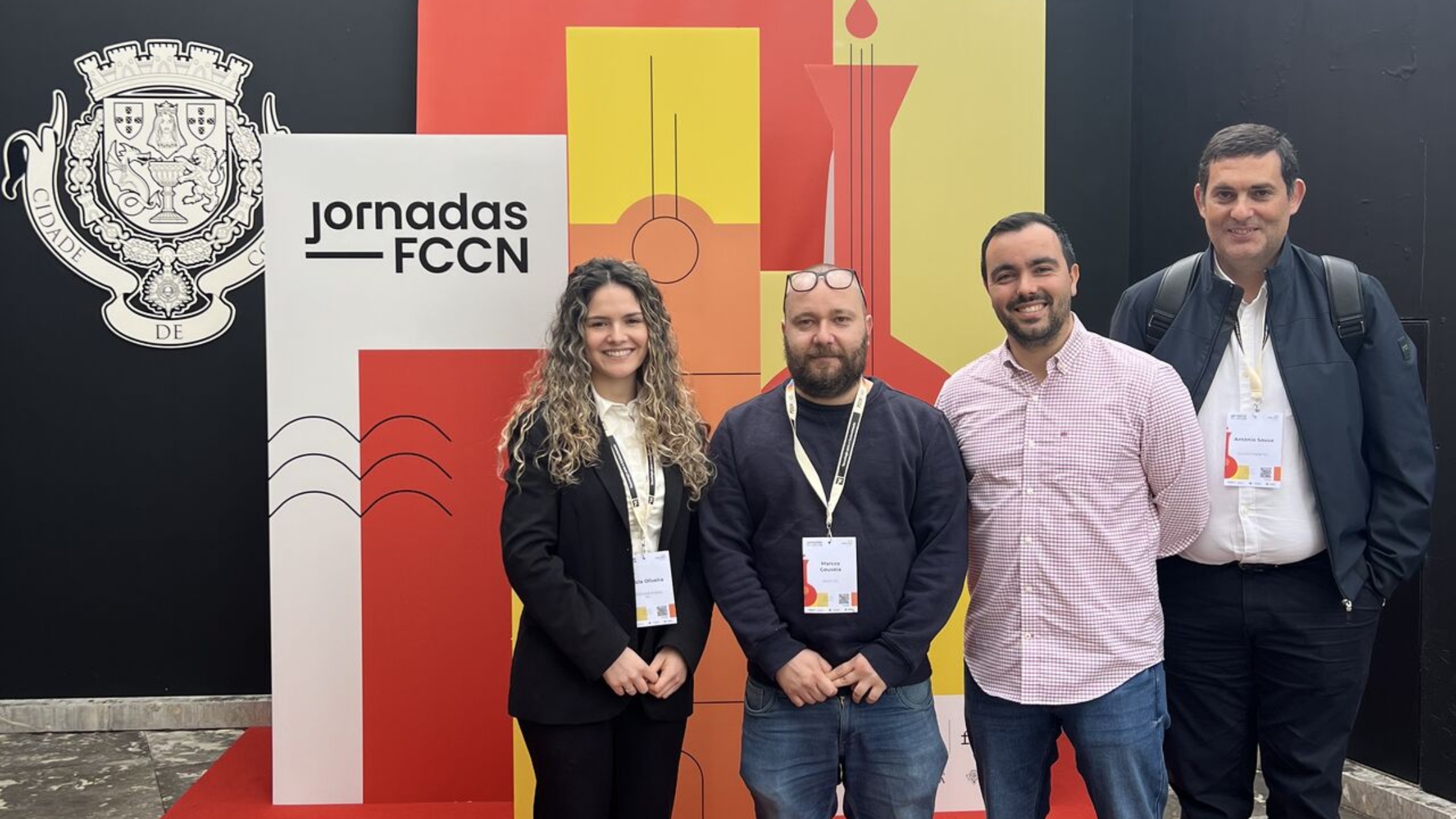 Four conference attendees standing together in front of a “Jornadas FCCN” event backdrop with colourful geometric panels and the Portuguese coat of arms displayed on the wall.
