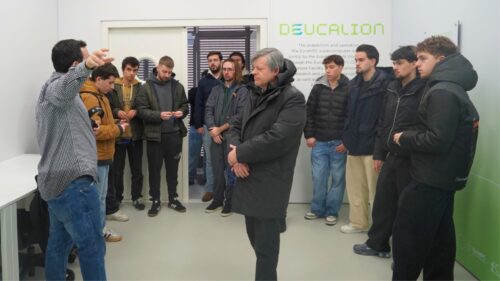 Um investigador fala para um grupo de quinze participantes durante uma reunião interna, gesticulando enquanto explica, com informações sobre o projeto Deucalion exibidas na parede atrás dele.