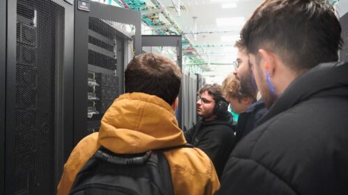 Dois visitantes do sexo masculino e um investigador do sexo masculino, usando proteção auditiva, examinam as unidades de refrigeração traseiras do supercomputador Deucalion, com grandes ventiladores e módulos de alimentação visíveis ao longo de uma fila de armários de computadores.