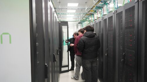 Um pequeno grupo de pessoas em pé entre filas de racks de servidores num centro de dados, com uma pessoa a explicar o equipamento dentro de um armário aberto.