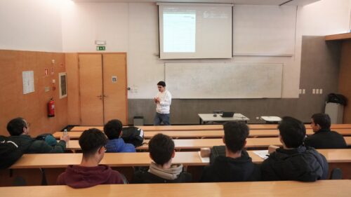 Investigador a apresentar para um grupo de estudantes numa sala de aula universitária, com slides projetados num ecrã.