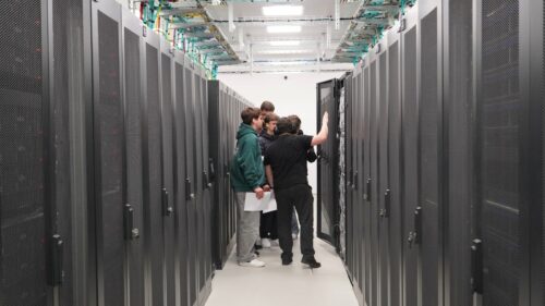 Grupo de estudantes entre as filas de racks de servidores num centro de dados, enquanto um investigador explica os componentes dentro de um armário aberto.