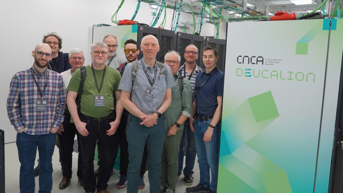 Grupo de participantes em pé no interior de um centro de dados, junto a racks de servidores e a um painel do sistema "Deucalion", durante uma visita às instalações de infraestrutura de HPC.