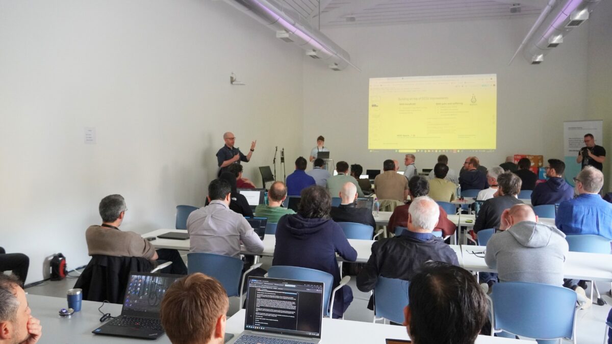 Público presente numa sessão de um workshop técnico, com os participantes sentados à volta de mesas a utilizar computadores portáteis, enquanto um orador apresenta slides na parte da frente da sala.