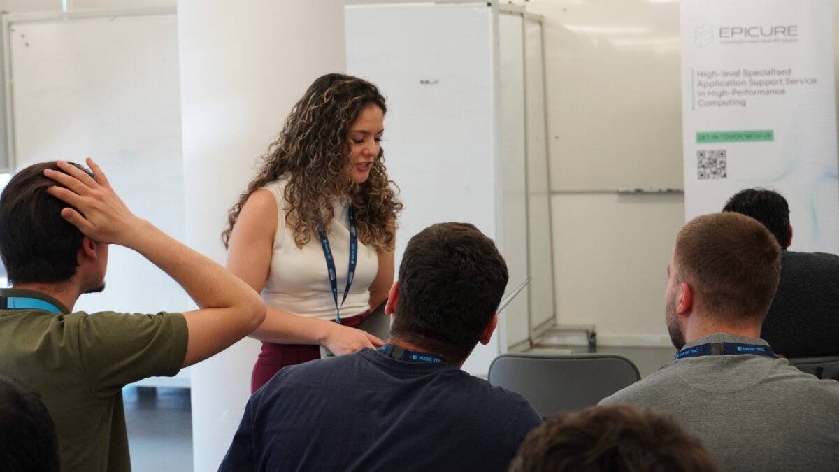 Mentor a dar suporte aos participantes de um workshop em HPC, a falar com um aluno com um computador portátil aberto enquanto outros participantes ouvem e interagem.