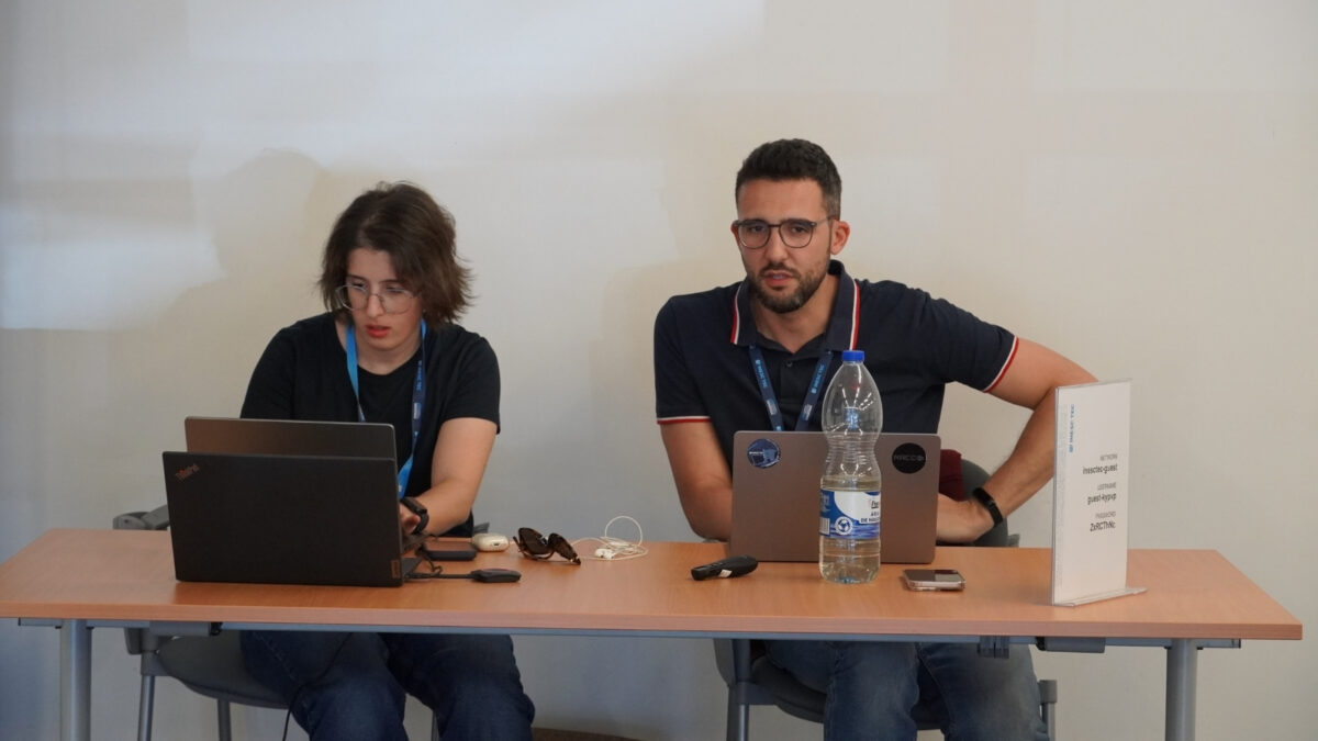Dois mentores do EPICURE Hackathon sentados numa mesa com computadores portáteis e materiais do evento em frente.