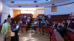 A speaker presents to an audience in a lecture hall, with attendees following the session from tiered seating