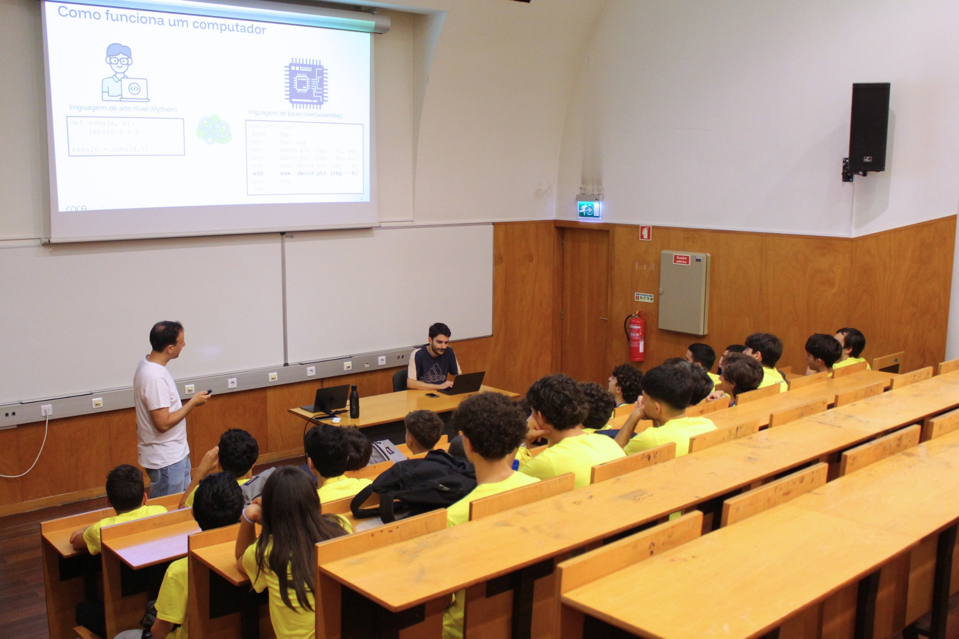 Um orador explica como funciona um computador a um grupo de estudantes que vestem t-shirts amarelas numa sala de aula da universidade.