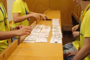Um grupo de alunos vestidos com t-shirts amarelas organiza cartas de jogar em filas ordenadas sobre uma secretária de madeira, durante uma atividade na sala de aula.