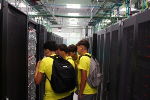 Um grupo de estudantes, vestidos com t-shirts amarelas e equipados com protetores auriculares, examina o equipamento entre as filas de armários de servidores durante uma visita ao centro de supercomputação do Deucalion.