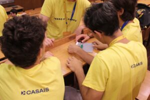 Um grupo de alunos vestidos com t-shirts amarelas trabalha em conjunto para montar peças de LEGO numa placa de base cinzenta durante uma atividade na sala de aula.