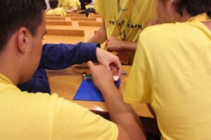 Os alunos, vestidos com t-shirts amarelas, trabalham em conjunto para montar peças de LEGO numa base azul durante uma atividade do HPC.